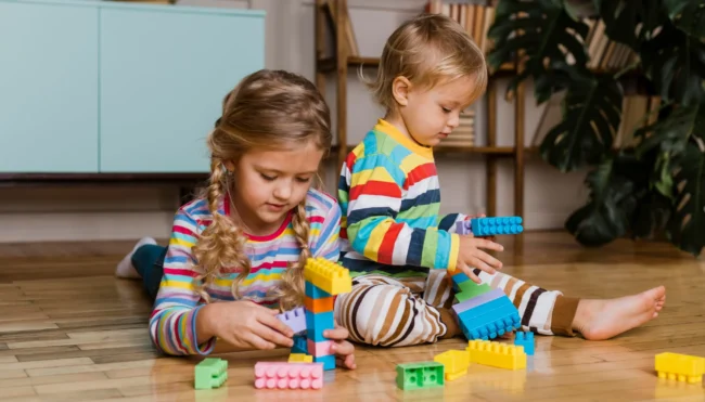 bloques-construccion-grandes-ninos-preescolar Niños pequeños jugando en el suelo con bloques de construcción de colores de tamaño grande para fomentar la creatividad.